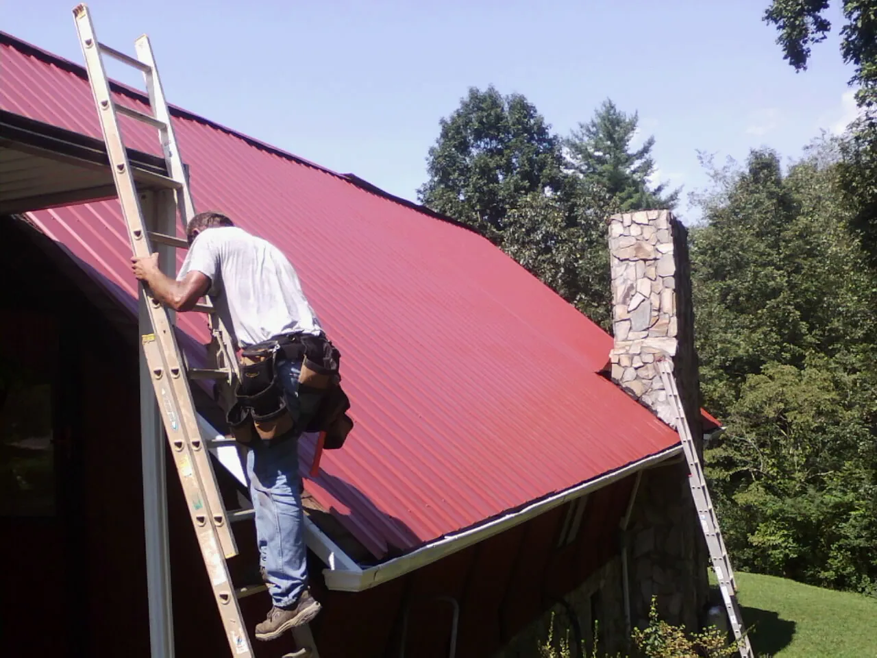 Roofer on a ladder beside a red metal roof and stone chimney — Soffit & Fascia Repair by Roofing Brothers in Southold